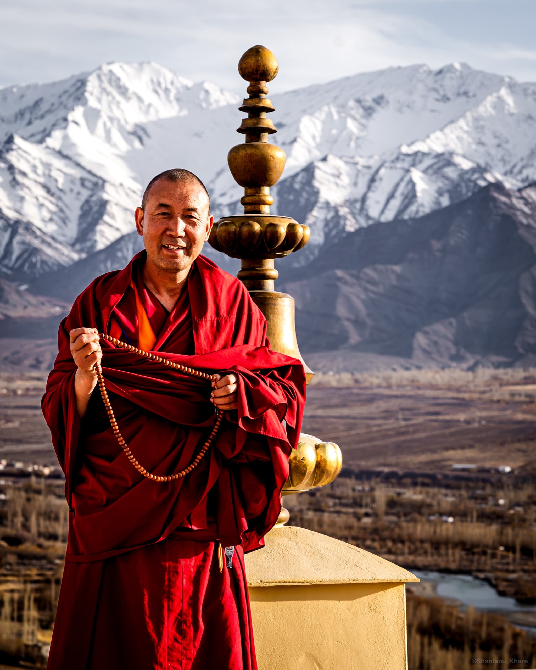 Documentary portrait photography — a Buddhist monk in red robes with prayer beads before snow peaks, Ladakh.