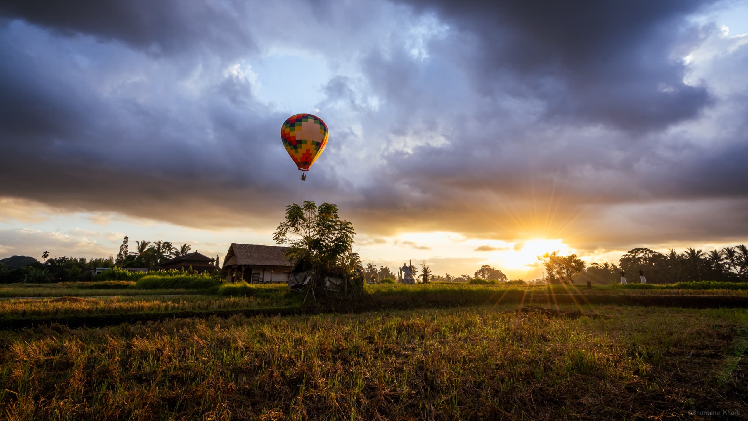 Landscape photography — a hot-air balloon drifting over rice fields as the sun breaks through storm clouds.