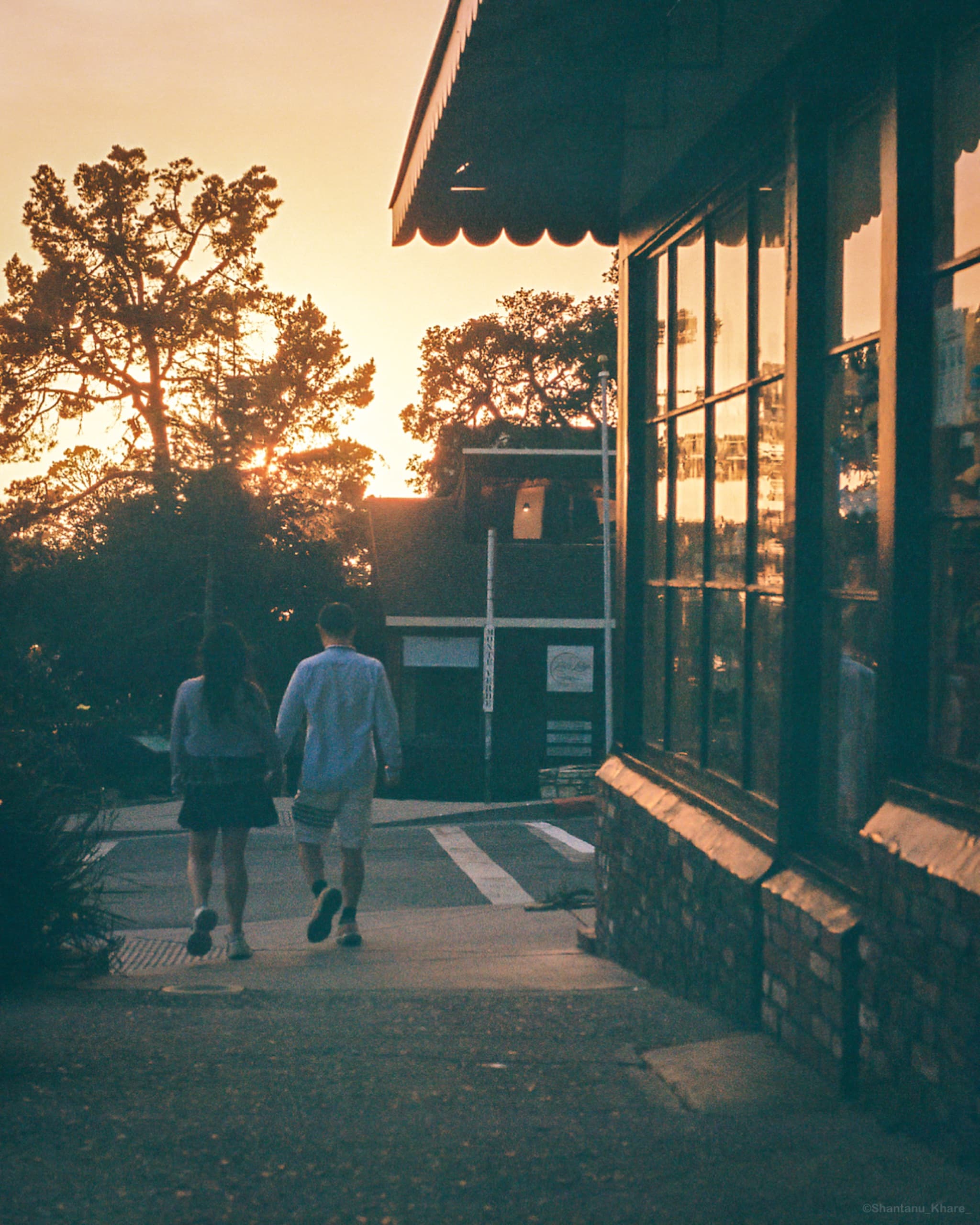 Film photography — a couple walking past a warm shopfront at sunset.