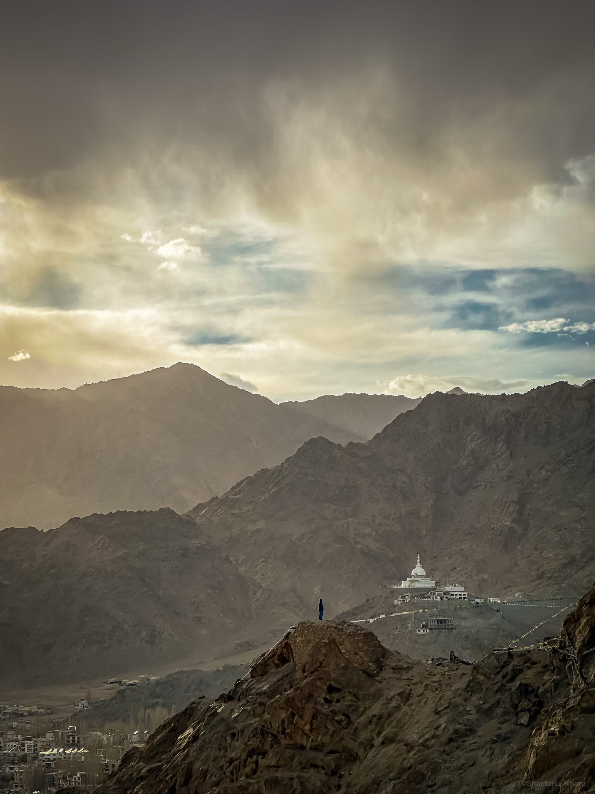 Landscape photography — a lone figure by a white stupa on a ridge as rain drifts over the mountains, Ladakh.