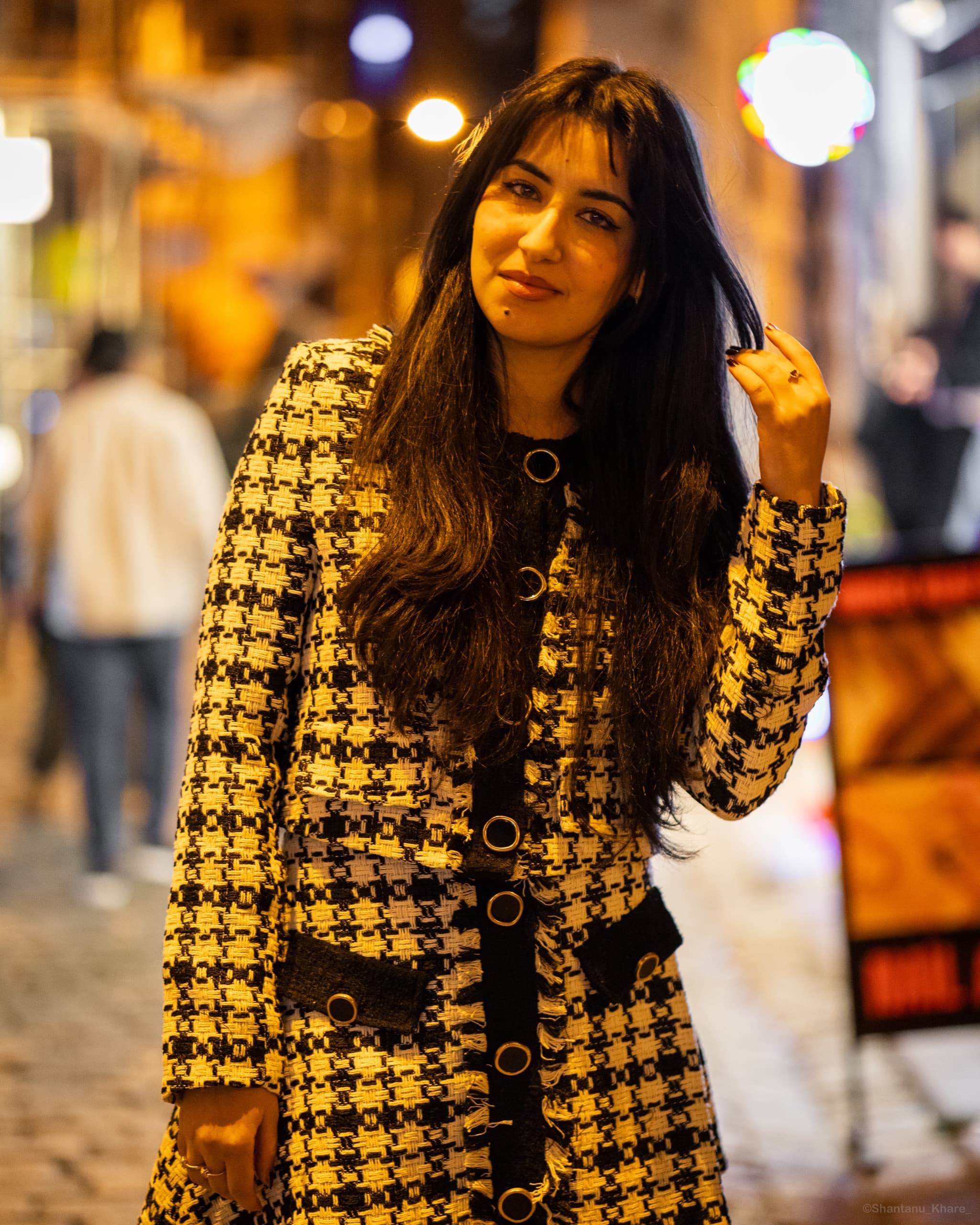 Street portrait photography — a woman in a houndstooth coat on a lamplit evening street.